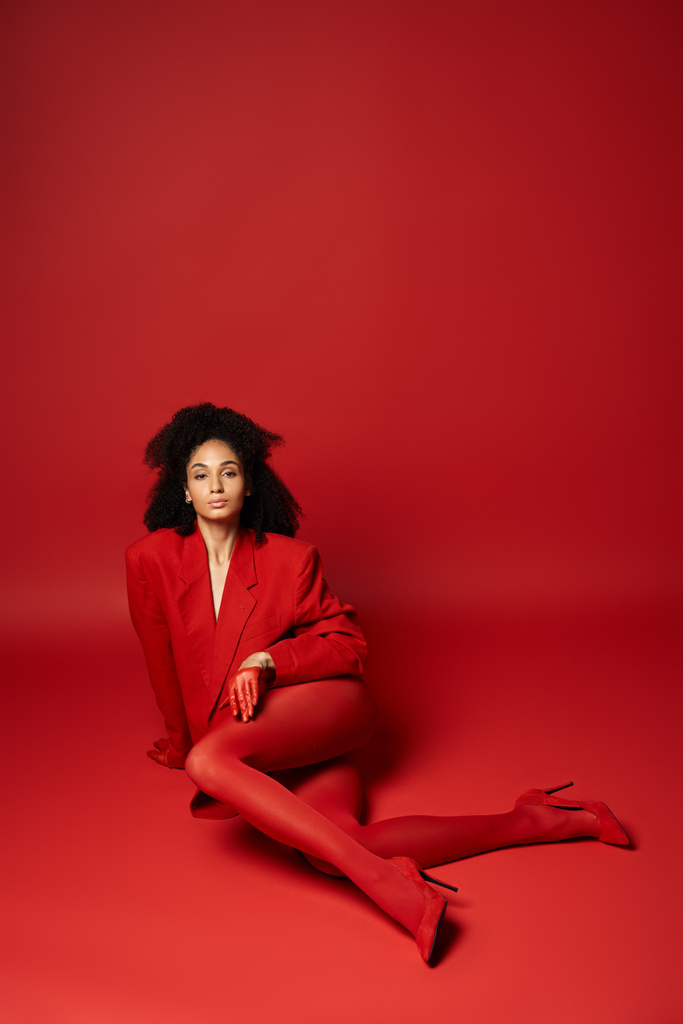 Uma jovem mulher senta-se graciosamente no chão, vestindo uma jaqueta vermelha brilhante e meia-calça contra um cenário vibrante em um estúdio. - Foto, Imagem