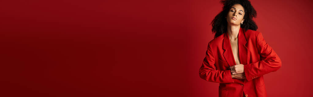 Μια νεαρή γυναίκα με ένα ζωηρό κόκκινο κοστούμι στέκεται με αυτοπεποίθηση μπροστά σε έναν τοίχο που ταιριάζει. - Φωτογραφία, εικόνα