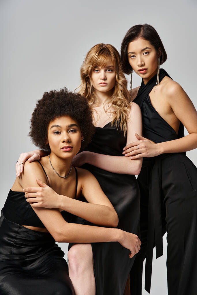 Drie vrouwen in zwarte jurken staan gracieus, tonen diversiteit en schoonheid met een grijze studio achtergrond. - Foto, afbeelding