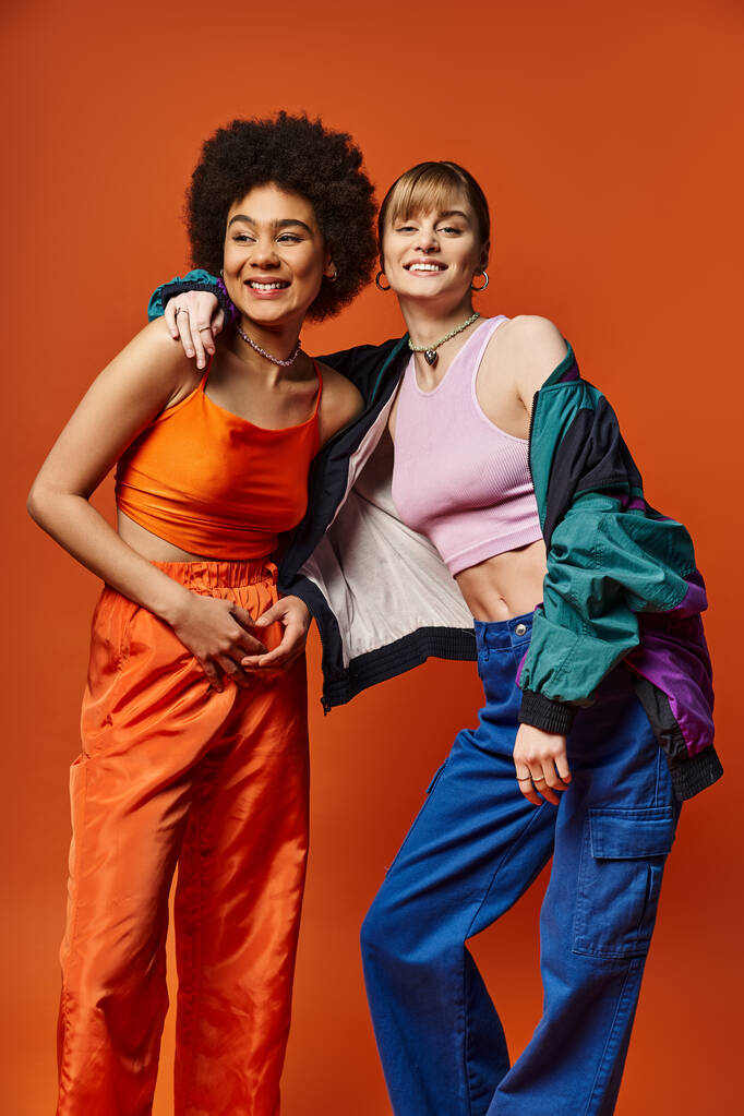 Duas belas mulheres de diferentes etnias estão lado a lado em um estúdio, sorrindo e posando para uma foto contra um fundo laranja. - Foto, Imagem