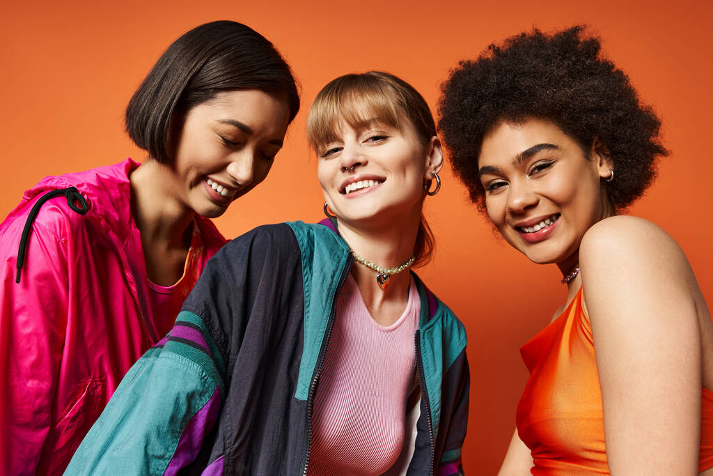 Tres hermosas mujeres de diferentes etnias de pie juntas frente a un telón de fondo naranja. - Foto, Imagen