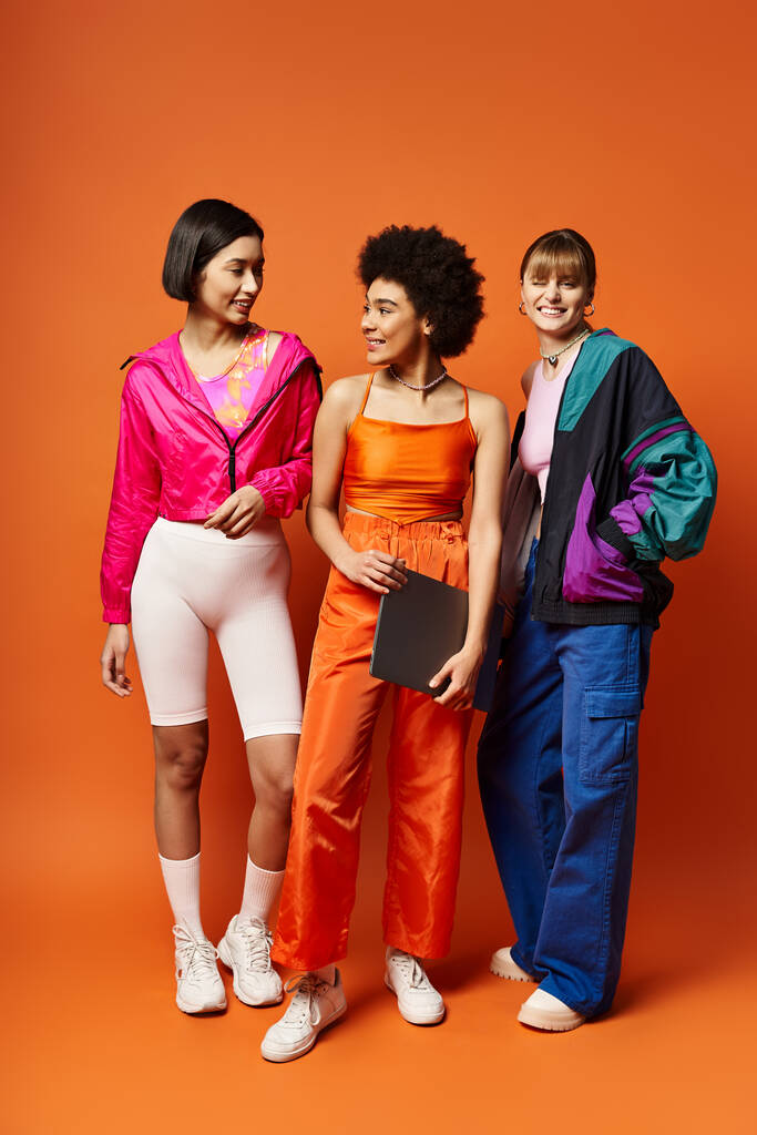 Tres hermosas mujeres de diferentes etnias de pie unidas frente a un fondo naranja. - Foto, Imagen