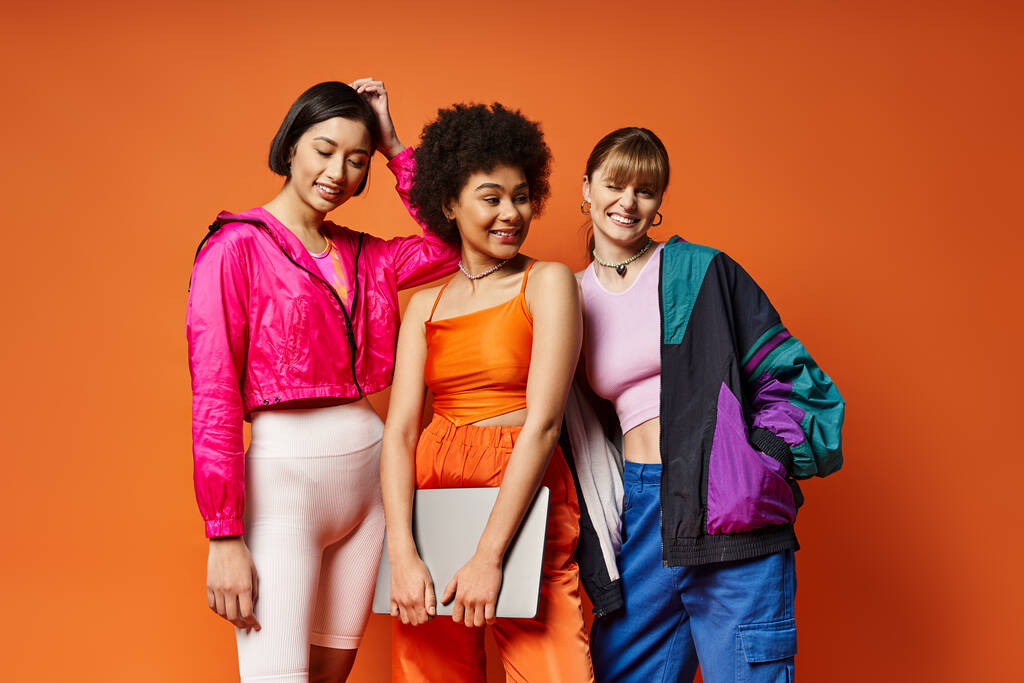 Three diverse women - Caucasian, Asian, African American - stand together against an orange studio backdrop, radiating beauty and unity. - Photo, Image