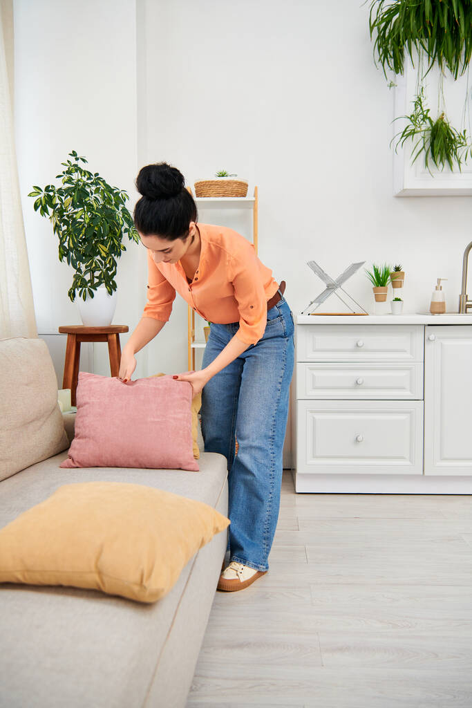 Una mujer elegante con atuendo casual coloca cuidadosamente una almohada decorativa en un sofá moderno, agregando un toque acogedor a su espacio de vida.. - Foto, Imagen