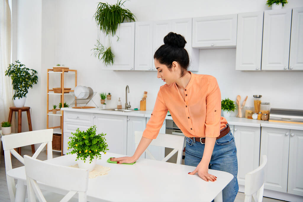 Une femme élégante en tenue décontractée essuie méticuleusement une table dans une cuisine maison, créant un espace lumineux et accueillant. - Photo, image