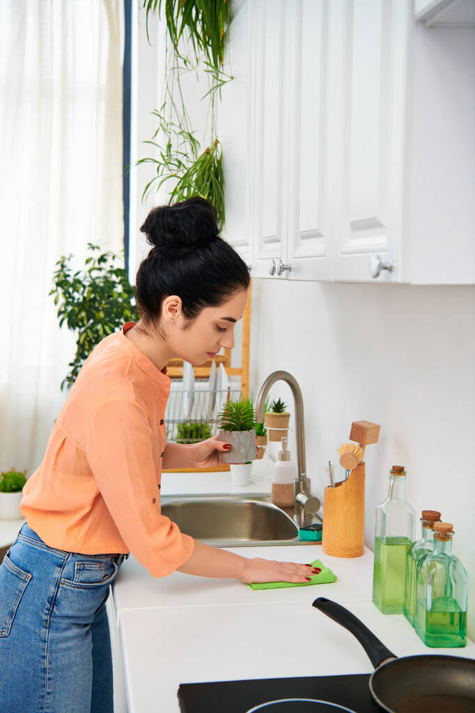Μια νεαρή γυναίκα με casual ενδυμασία καθαρίζει ένα νεροχύτη από ανοξείδωτο χάλυβα σε μια ζεστή κουζίνα, που περιβάλλεται από σαπούνια και είδη καθαρισμού. - Φωτογραφία, εικόνα