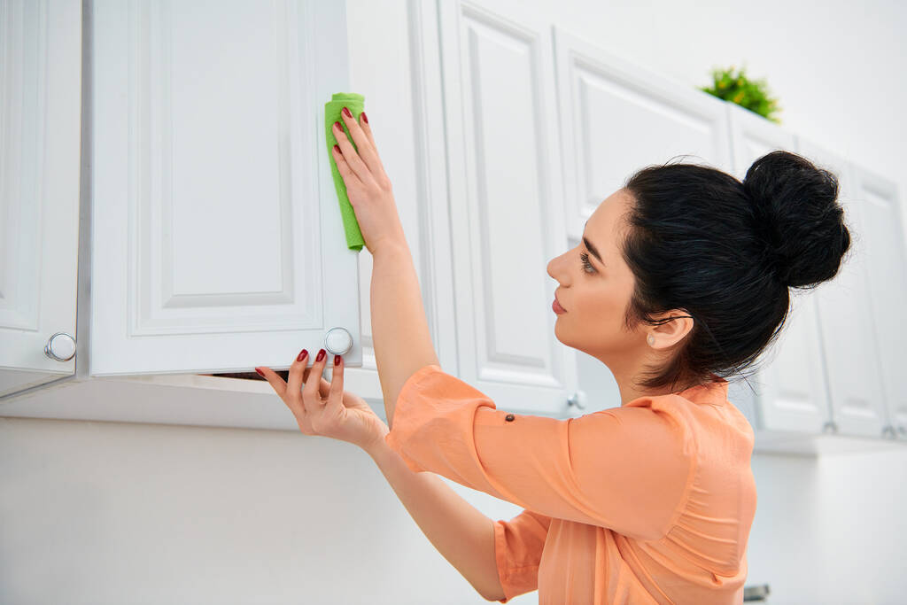 Una mujer con atuendo casual limpia meticulosamente una cocina usando un trapo verde, asegurando que cada superficie brille con un brillo. - Foto, Imagen
