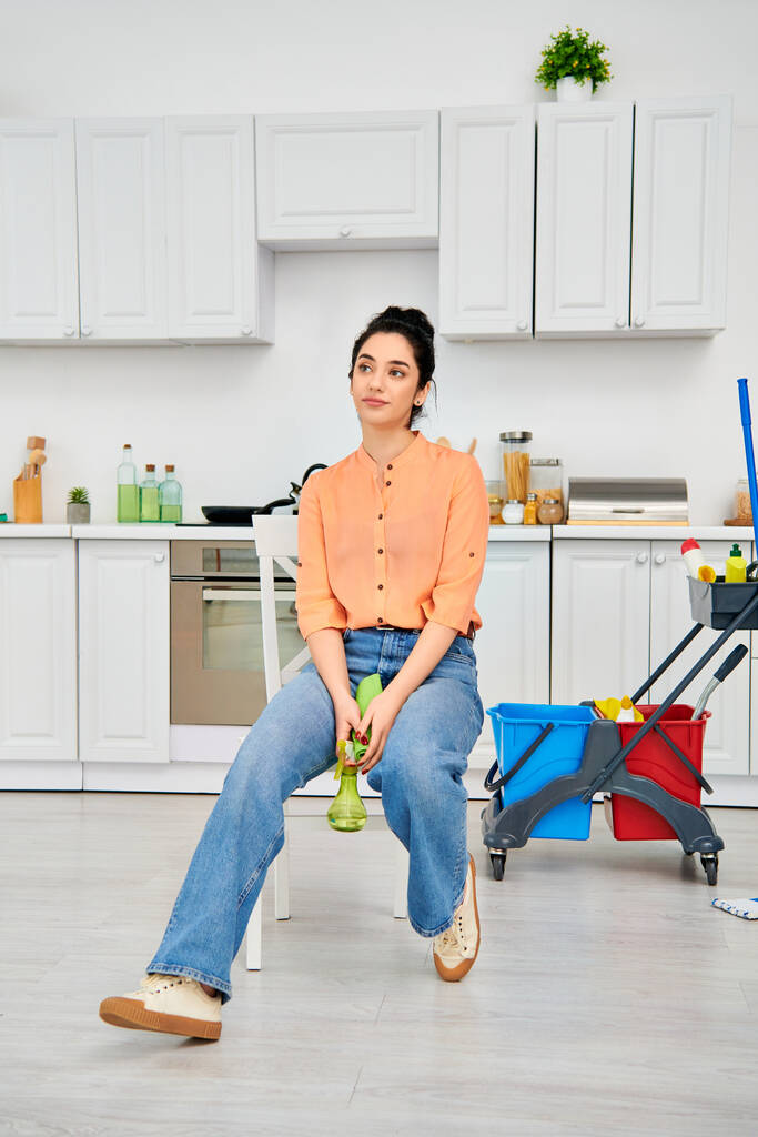 Gündelik giysiler içinde şık bir kadın arka planda pırıl pırıl temiz bir mutfak olan bir sandalyeye oturur.. - Fotoğraf, Görsel