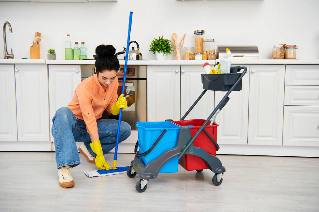 Μια κομψή γυναίκα με casual ενδυμασία καθαρίζει με χάρη το πάτωμα με μια σφουγγαρίστρα και κουβά, φέρνοντας μια πινελιά κομψότητας στις δουλειές του σπιτιού της. - Φωτογραφία, εικόνα