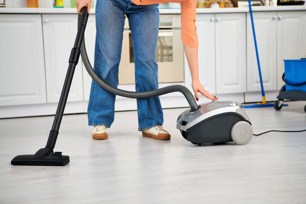A stylish woman in casual attire gracefully vacuums the kitchen floor. - Photo, Image