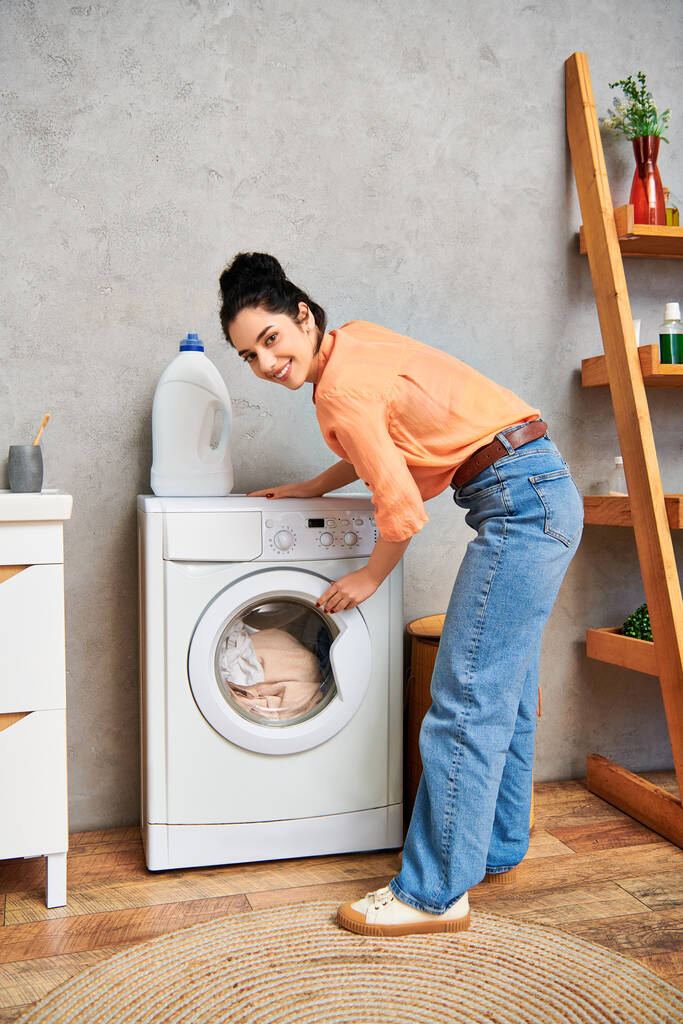 Uma mulher elegante em trajes casuais fica ao lado de uma máquina de lavar roupa, focada na limpeza de suas roupas em casa. - Foto, Imagem