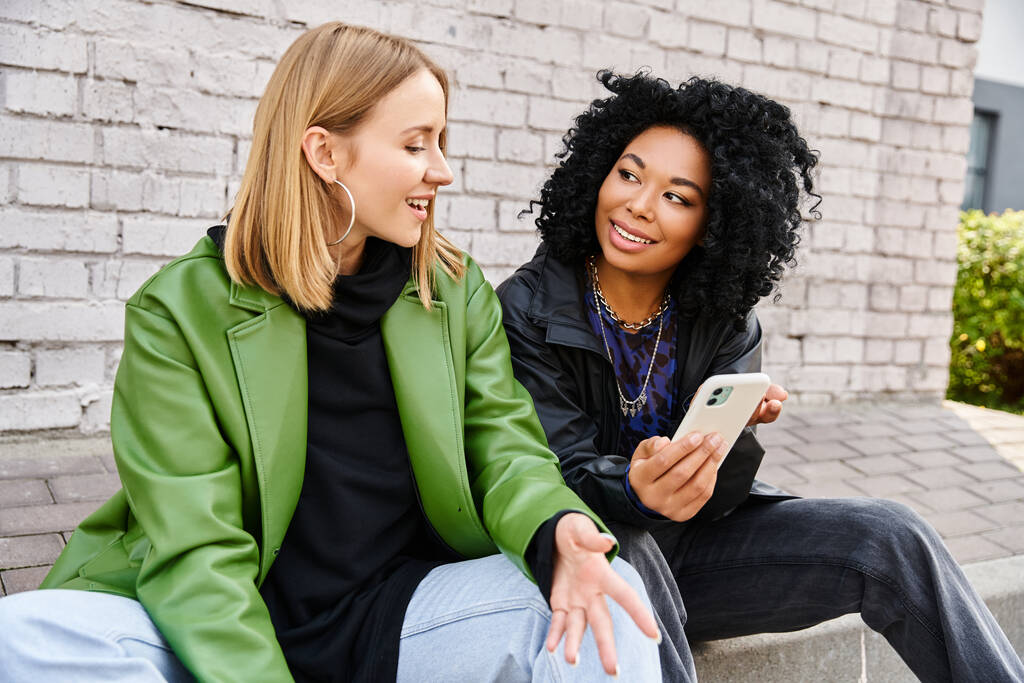 Two diverse women in casual attire sit on a bench, engrossed in a cell phone. - Photo, Image