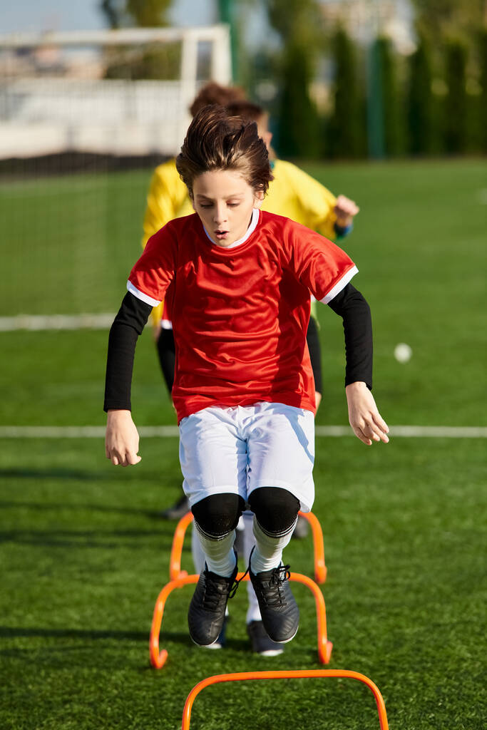 Ένα νεαρό αγόρι κλωτσάει μια μπάλα ποδοσφαίρου στο γήπεδο του πράσινου ποδοσφαίρου, δείχνοντας το πάθος και τις ικανότητές του στο άθλημα. Η συγκέντρωση και η αποφασιστικότητά του είναι προφανείς καθώς επικεντρώνεται στην τελειοποίηση της τεχνικής του.. - Φωτογραφία, εικόνα