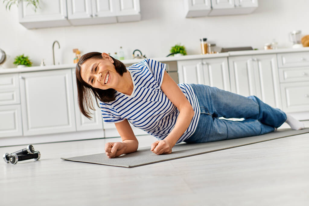 Una hermosa mujer madura en acogedora ropa de casa practica yoga en una alfombra en su cocina. - Foto, imagen