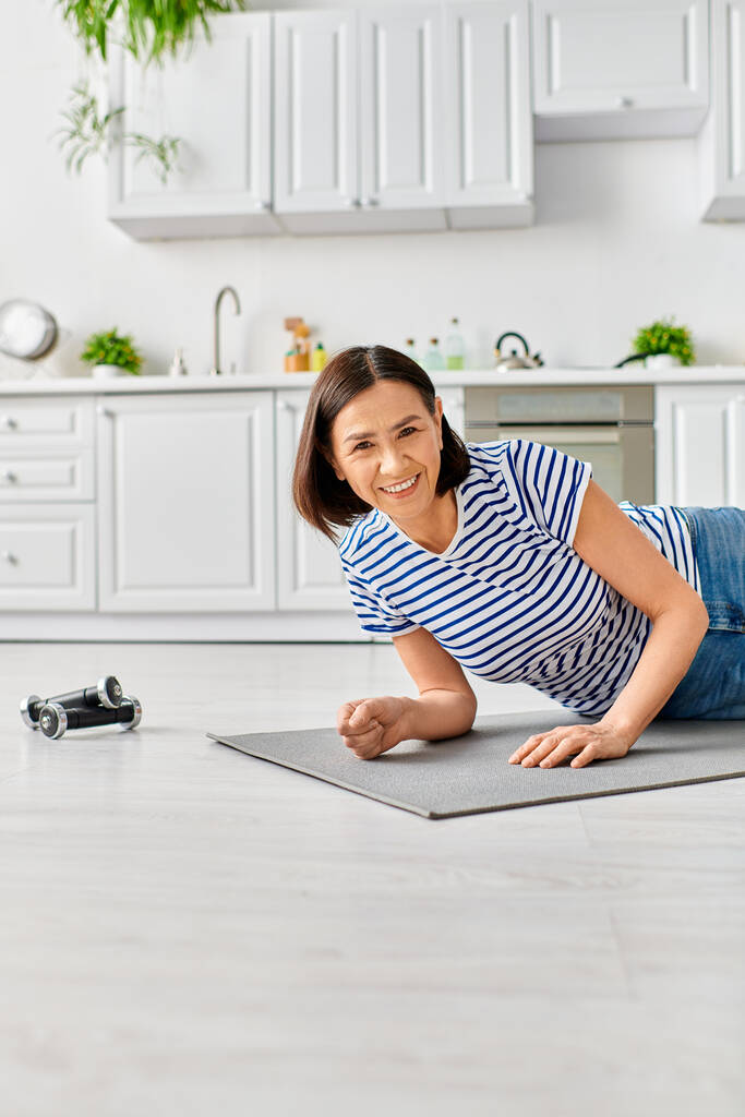 Dojrzała kobieta w homewear ćwiczy jogę na macie w słonecznej kuchni. - Zdjęcie, obraz