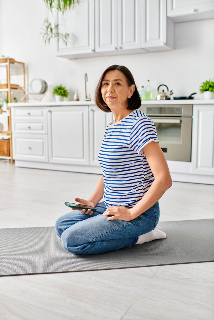 Une belle femme mature en vêtements confortables effectue du yoga sur un tapis dans sa cuisine. - Photo, image