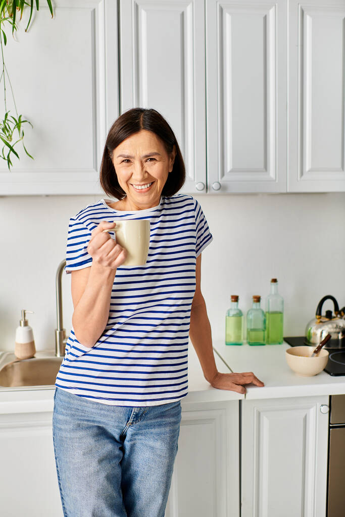 Uma mulher em roupa de casa aconchegante fica em uma cozinha, segurando um copo. - Foto, Imagem