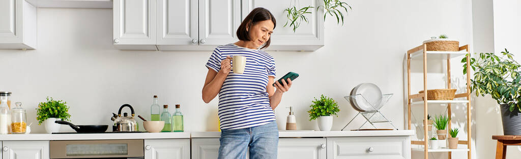 Una mujer madura en ropa de casa acogedora se encuentra en una cocina, sosteniendo un teléfono celular. - Foto, imagen