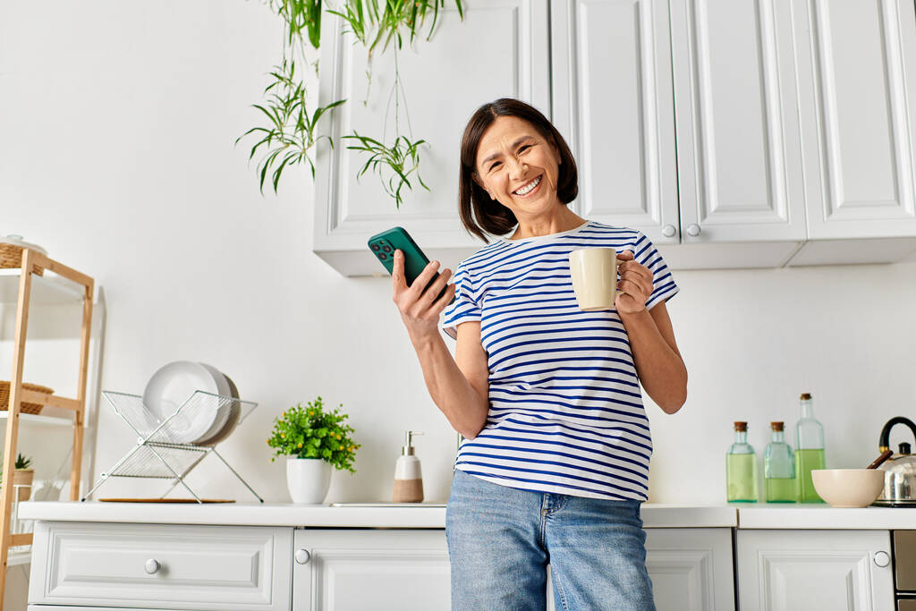 Femme dans des vêtements confortables tenant tasse et téléphone cellulaire dans la cuisine. - Photo, image