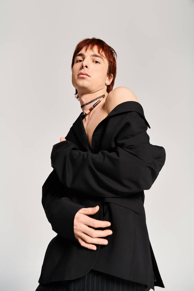 Un joven elegante con el pelo rojo posa con confianza en una chaqueta negra sobre un fondo gris de estudio. - Foto, imagen