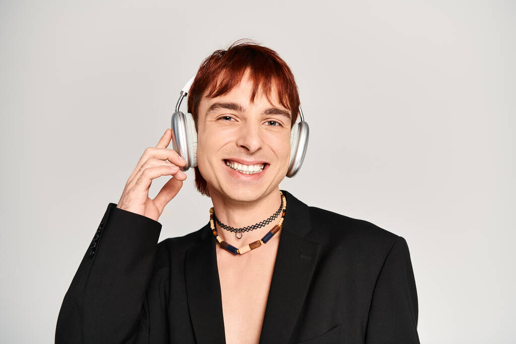 Ένας κομψός νεαρός με κόκκινα μαλλιά που ακούει μουσική μέσα από ακουστικά σε ένα στούντιο με γκρι φόντο. - Φωτογραφία, εικόνα