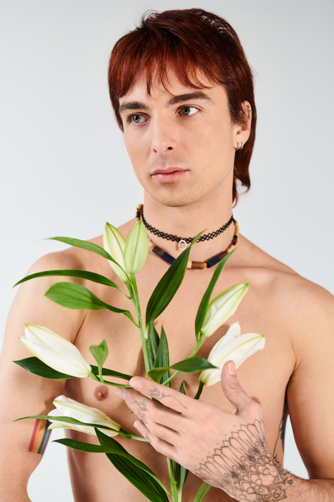 Ένας γυμνός νεαρός άνδρας σε ένα στούντιο κρατά ένα ζωντανό φυτό στο χέρι του πάνω σε ένα γκρι φόντο. - Φωτογραφία, εικόνα
