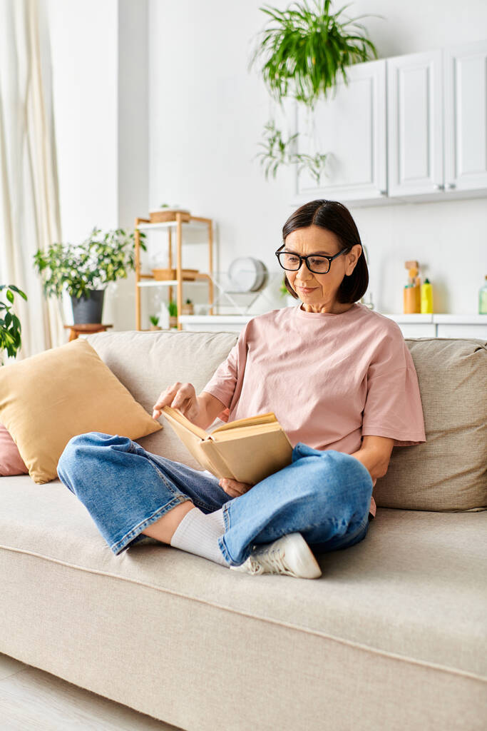 Egy érett nő kényelmes otthonban ül a kanapén, teljesen elmerülve egy könyv olvasásában.. - Fotó, kép