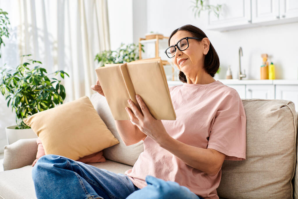 Зрелая женщина в домашней одежде, сидящая на диване, поглощенная книгой. - Фото, изображение