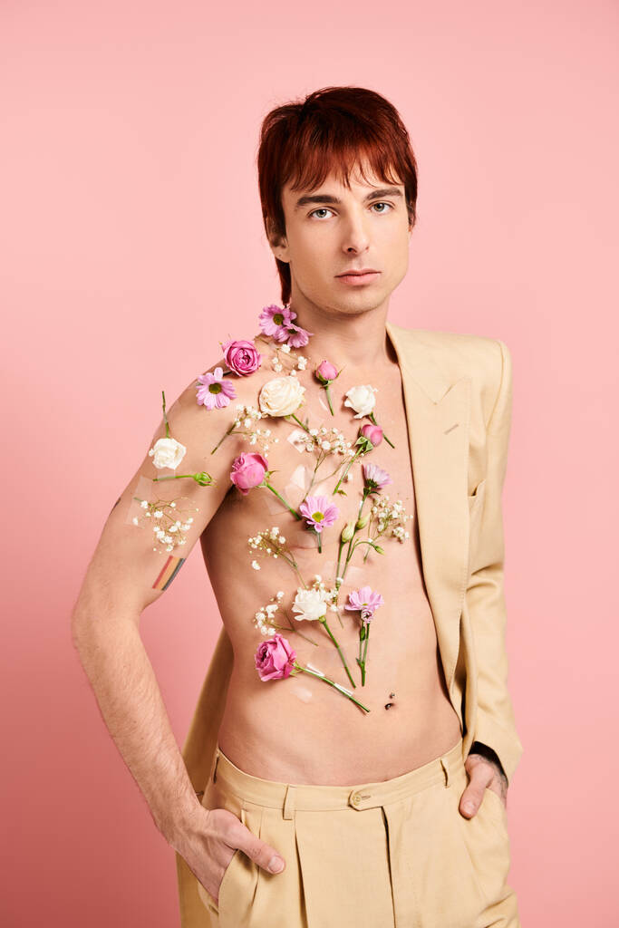 Ένας νεαρός άνδρας χωρίς μπλούζα ποζάρει με πολύχρωμα λουλούδια στολίζοντας το στήθος του σε ένα ροζ φόντο στούντιο. - Φωτογραφία, εικόνα