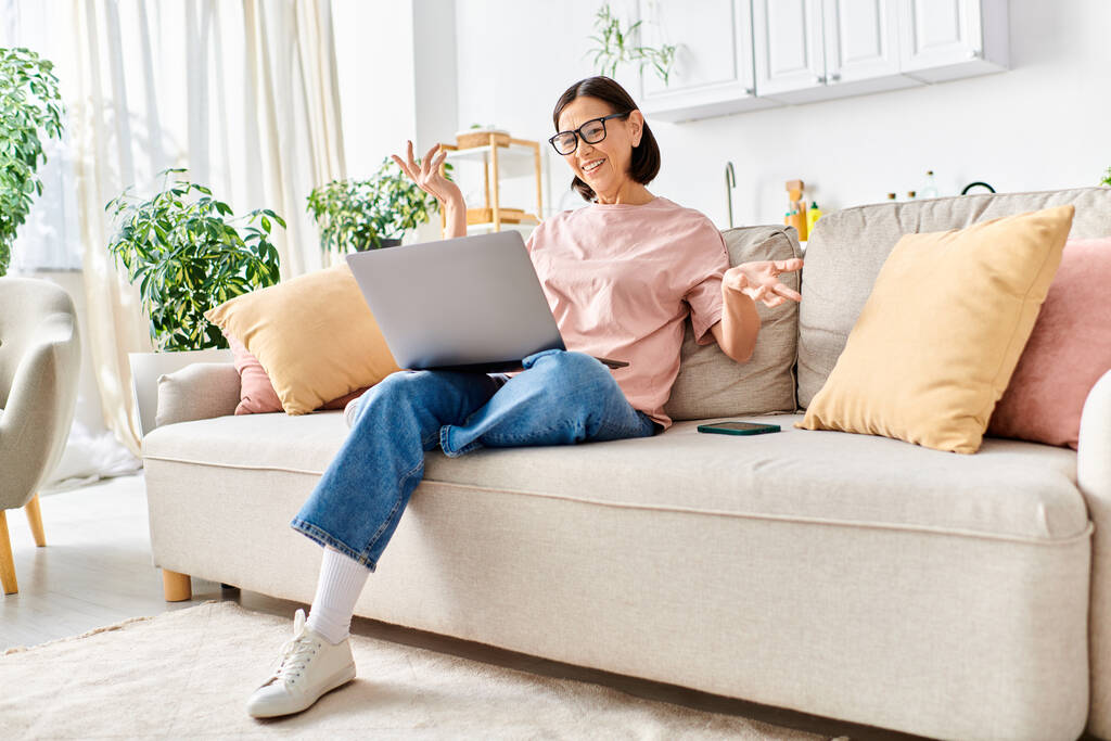 Uma mulher madura, em roupa de casa aconchegante, senta-se em um sofá usando um laptop. - Foto, Imagem