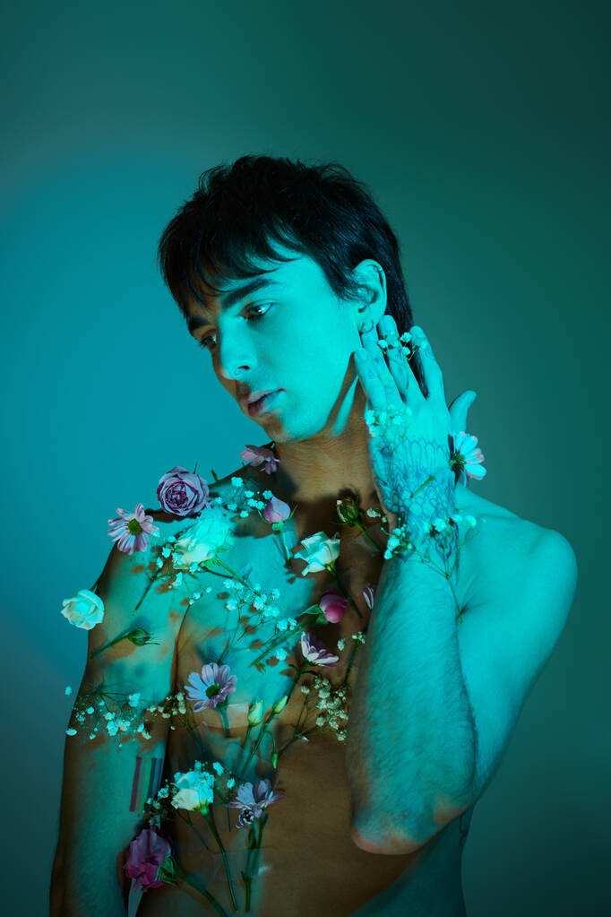 Tyylikäs nuori mies poseeraa kukkien kanssa kehonsa ympärillä studiossa, jossa on sininen valo. - Valokuva, kuva