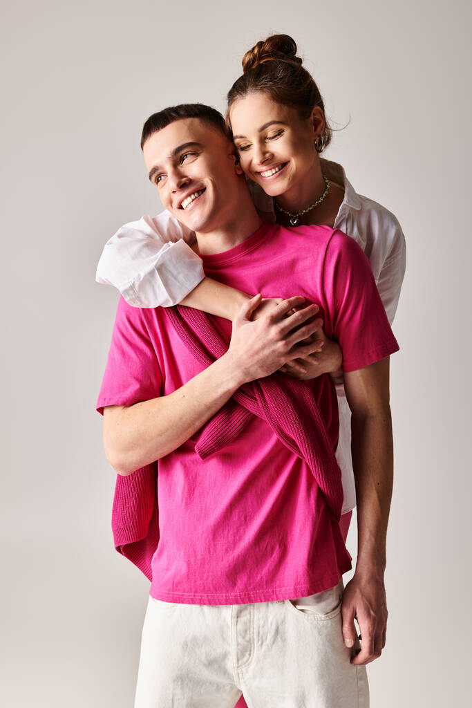 Ένας άντρας αγκαλιάζει στοργικά μια γυναίκα φορώντας ένα ροζ πουκάμισο σε ένα κομψό στούντιο με γκρι φόντο. - Φωτογραφία, εικόνα