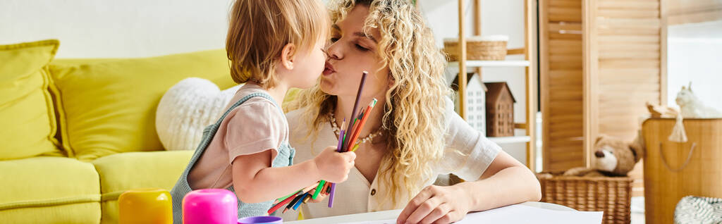 Une mère aux cheveux bouclés embrasse amoureusement sa petite fille sur la joue dans une maison confortable en utilisant la méthode d'éducation Montessori. - Photo, image