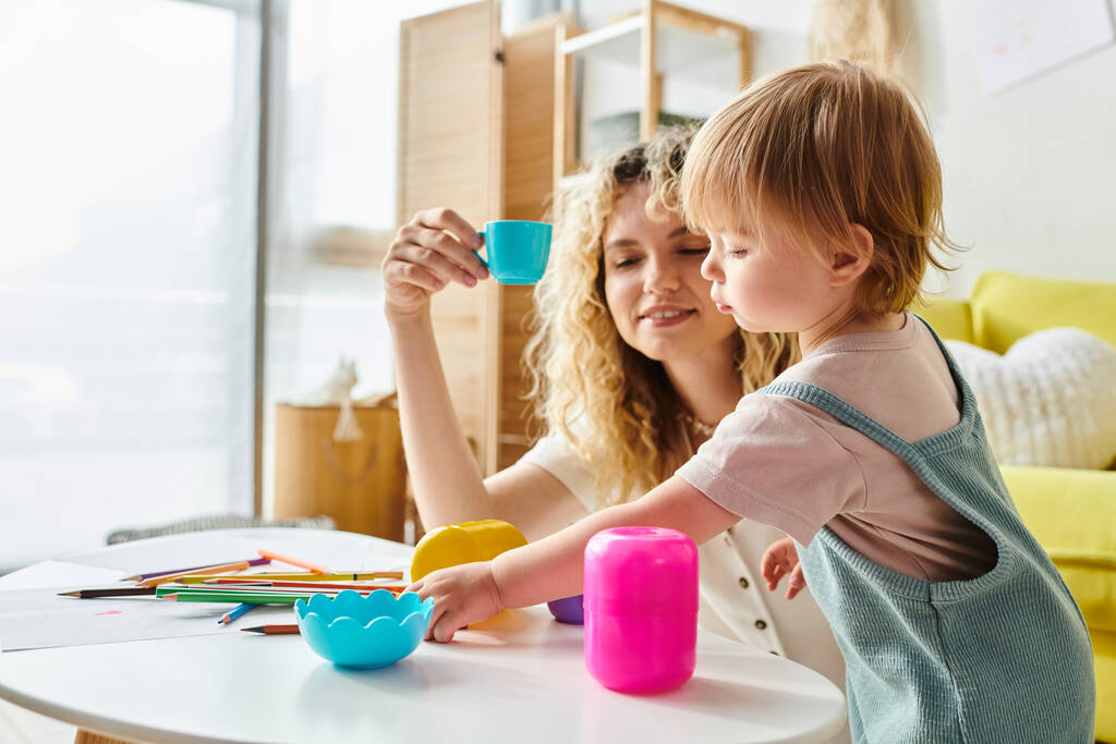Een krullende moeder en haar peuterdochter gaan speels op ontdekkingstocht met kleurrijke bekers thuis, waarbij ze de Montessori methode omarmen. - Foto, afbeelding