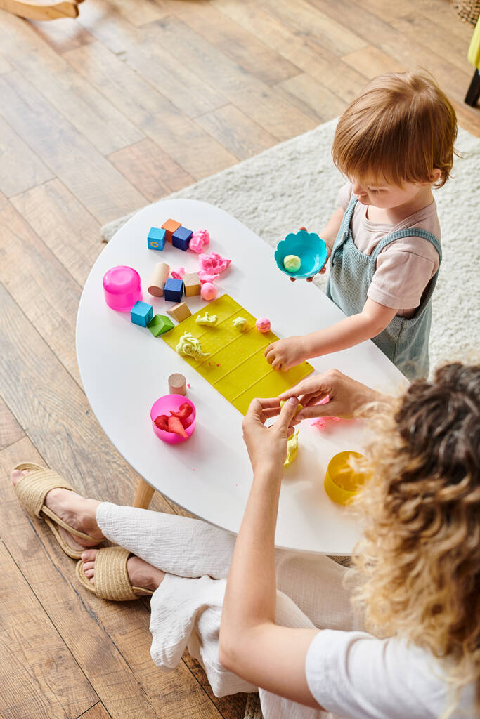 Μια σγουρή μητέρα και η κόρη της ασχολούνται με το παιχνίδι με παιχνίδια σε ένα τραπέζι χρησιμοποιώντας τη μέθοδο της εκπαίδευσης Montessori. - Φωτογραφία, εικόνα