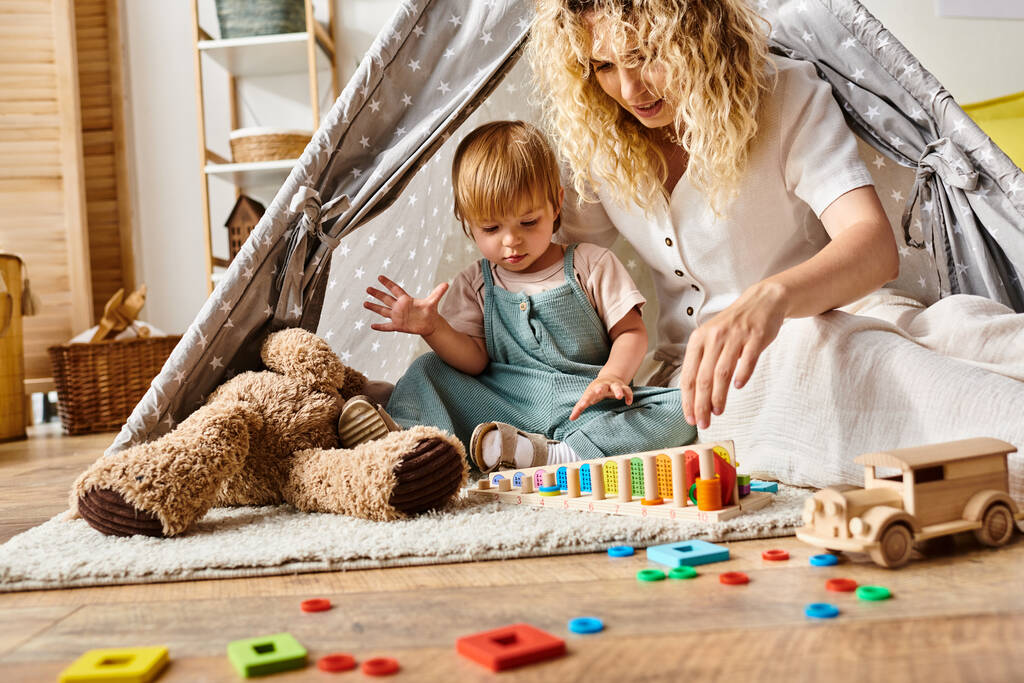 Кучерявая мать и маленькая дочь участвуют в творческой игре в красочной игровой палатке в уютной домашней обстановке. - Фото, изображение