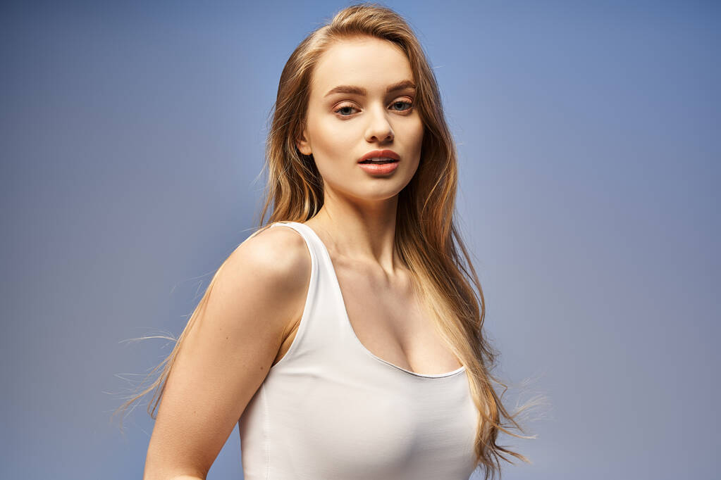 Μια νεαρή ξανθιά γυναίκα με λαμπερό χαμόγελο ποζάρει με χάρη σε ένα λευκό μπλουζάκι, αποπνέοντας ομορφιά και αυτοπεποίθηση σε ένα στούντιο.. - Φωτογραφία, εικόνα