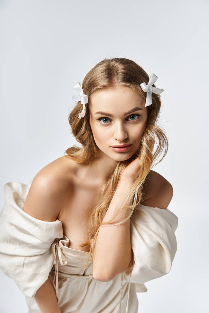 Oszałamiająca młoda kobieta z blond włosami z wdziękiem pozuje w płynącej białej sukience w studio. - Zdjęcie, obraz