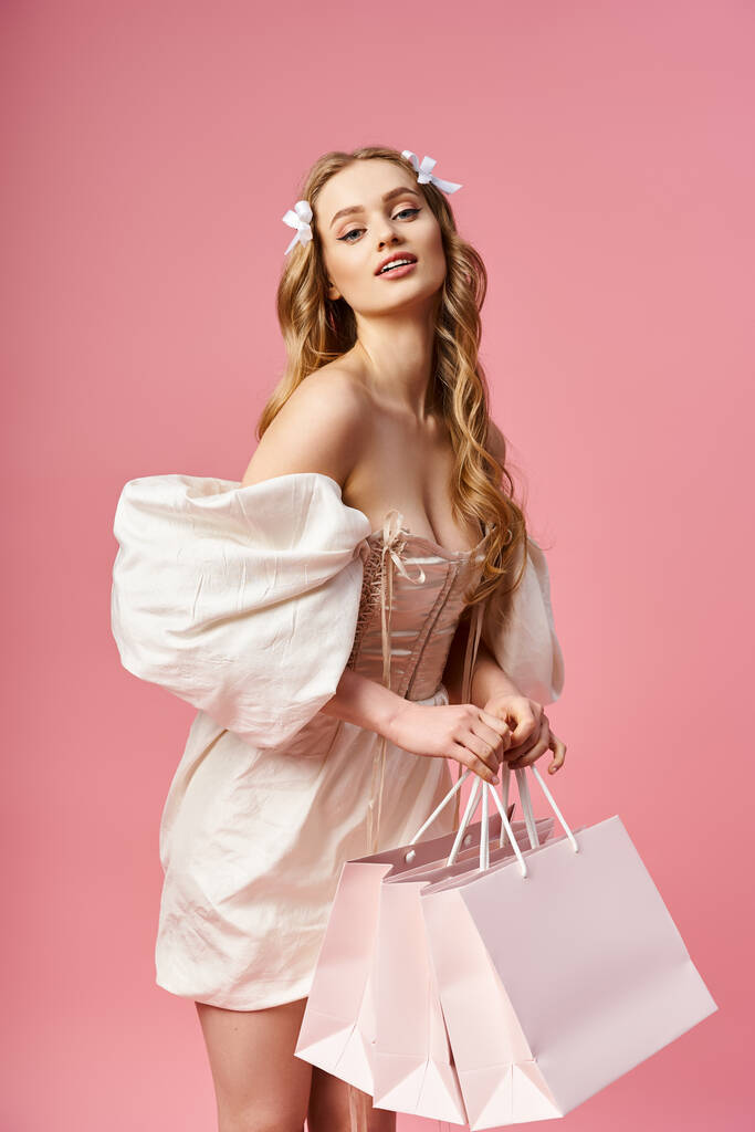 白いドレスの若い金髪の女性は優雅にスタジオの設定でカラフルなショッピングバッグを保持しています. - 写真・画像