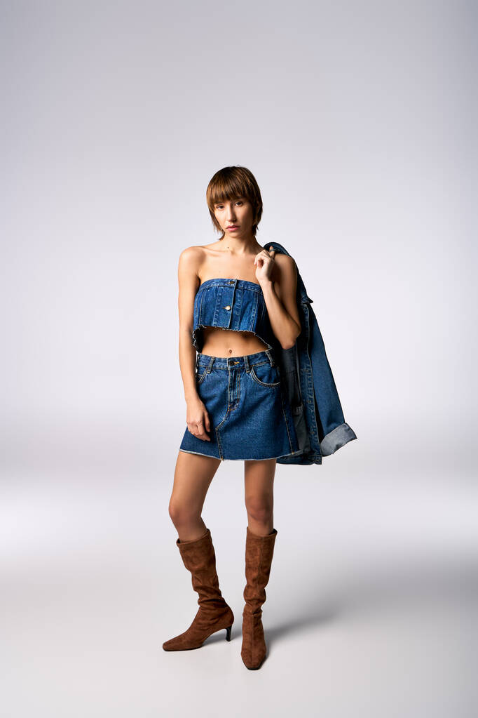 Eine selbstbewusste junge Frau mit kurzen Haaren posiert in Jeanskleid und Stiefeln für ein Mode-Shooting in einem Studio. - Foto, Bild