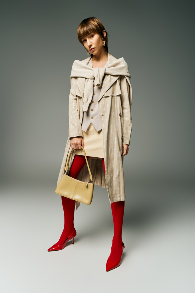 A young woman with short hair, wearing a trench coat, confidently holds a purse in a captivating and mysterious pose. - Photo, Image