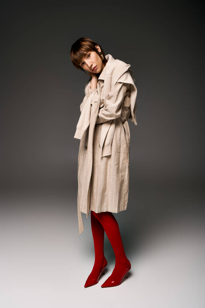 Uma jovem mulher com cabelo curto de pé em um casaco de trincheira e meias vermelhas em um ambiente de estúdio. - Foto, Imagem