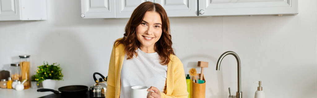 Middle-aged woman standing in kitchen, holding cup. - Photo, Image