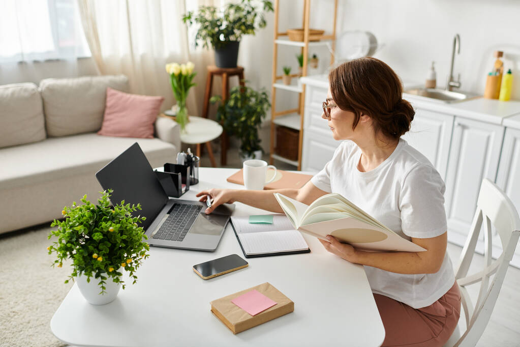 Kobieta w średnim wieku pochłonięta pracą, laptopem i książką na stole. - Zdjęcie, obraz