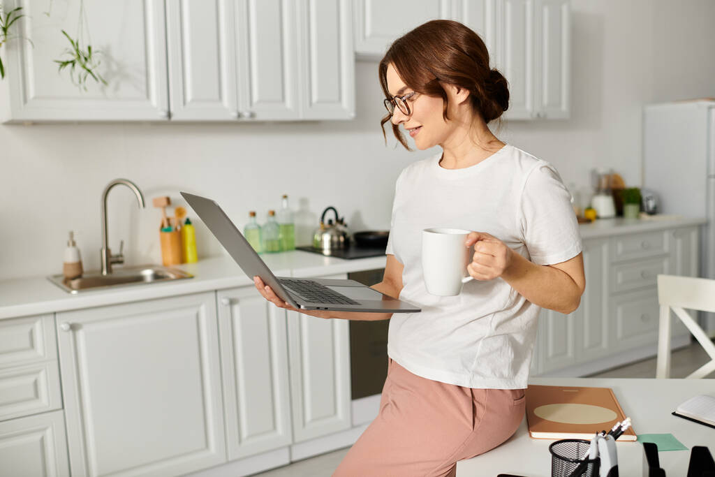 Kobieta w średnim wieku relaksuje się w domu, trzyma filiżankę kawy i pracuje na laptopie. - Zdjęcie, obraz