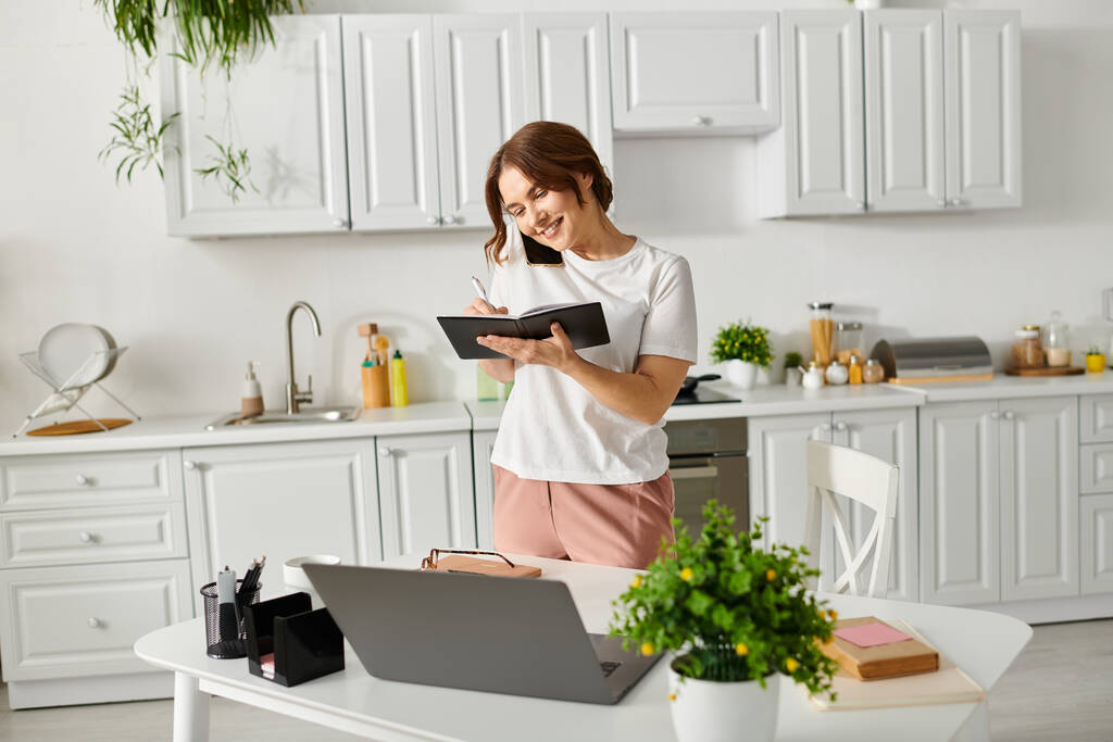 Жінка середнього віку поглинута читанням книги, стоячи на кухні. - Фото, зображення