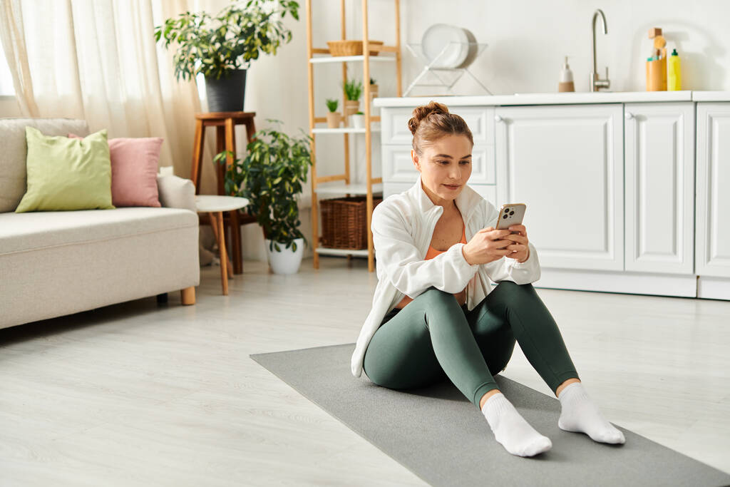 Femme d'âge moyen sur tapis de yoga, absorbé dans le smartphone. - Photo, image