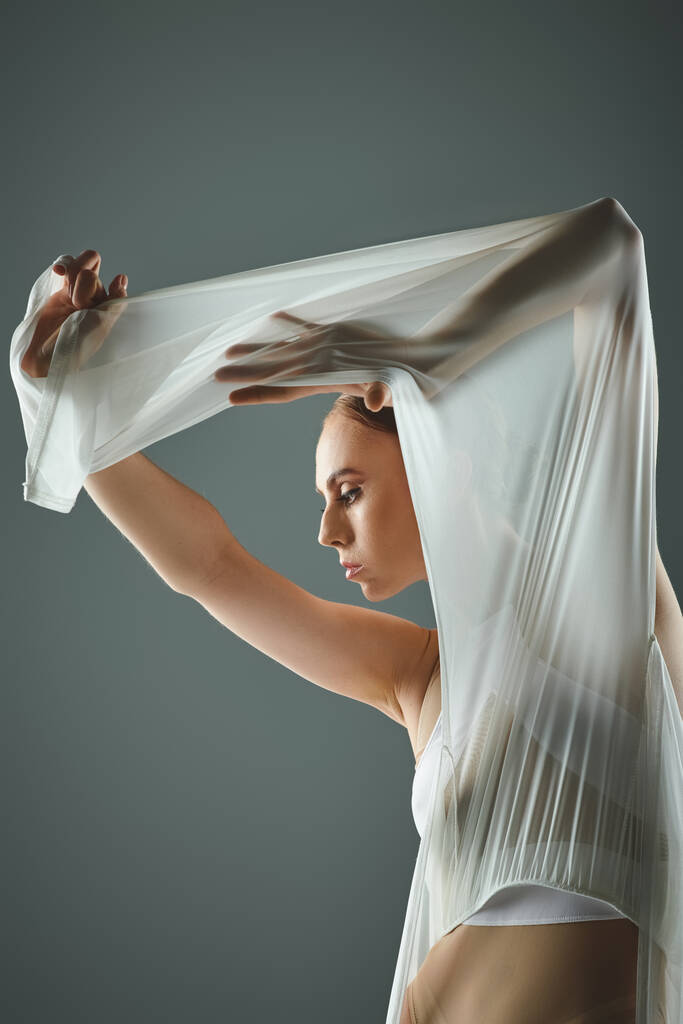 Een jonge ballerina in een witte jurk houdt haar sluier sierlijk over haar hoofd. - Foto, afbeelding