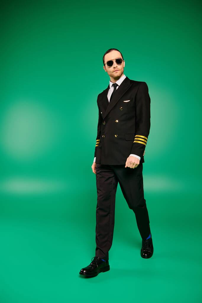 Ένας κομψός πιλότος με μαύρο κοστούμι και γυαλιά ηλίου στέκεται με αυτοπεποίθηση σε ένα φωτεινό πράσινο φόντο. - Φωτογραφία, εικόνα