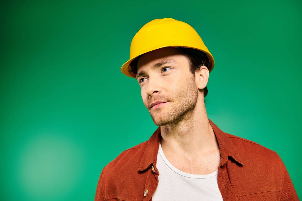 均一で堅い帽子のハンサムな労働者は鮮やかな緑の背景に対して自信を持って立っています. - 写真・画像